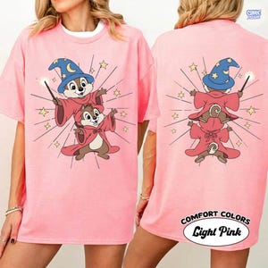 Puede incluir: Camiseta rosa claro con una ilustraci&oacute;n de dibujos animados de dos ardillas vestidas de magos, con un sombrero azul y una varita. La parte trasera de la camiseta tambi&eacute;n tiene el mismo dise&ntilde;o. El texto "COMFORT COLORS Light Pink" est&aacute; impreso en la parte inferior.