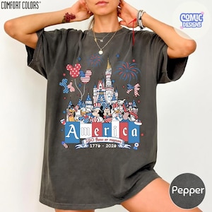 May include: A charcoal grey short-sleeve t-shirt featuring a graphic of a castle, fireworks, and cartoon characters in red, white, and blue. The text reads America, 250 Years of Freedom, and 1776-2026. The shirt has a Comfort Colors brand label.