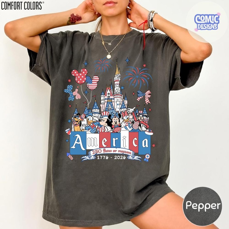 May include: A charcoal grey short-sleeve t-shirt featuring a graphic of a castle, fireworks, and cartoon characters in red, white, and blue. The text reads America, 250 Years of Freedom, and 1776-2026. The shirt has a Comfort Colors brand label.