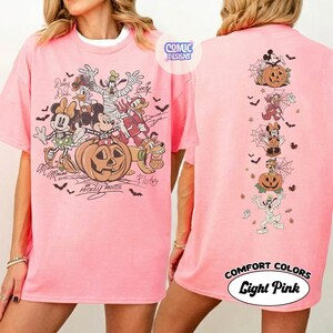 Op de afbeelding: Een lichtroze t-shirt met een grote Halloween-afbeelding op de voorkant met Mickey Mouse, Minnie Mouse, Donald Duck, Daisy Duck, Goofy en Pluto in kostuums. De achterkant toont een verticale kolom van dezelfde personages met pompoenen.