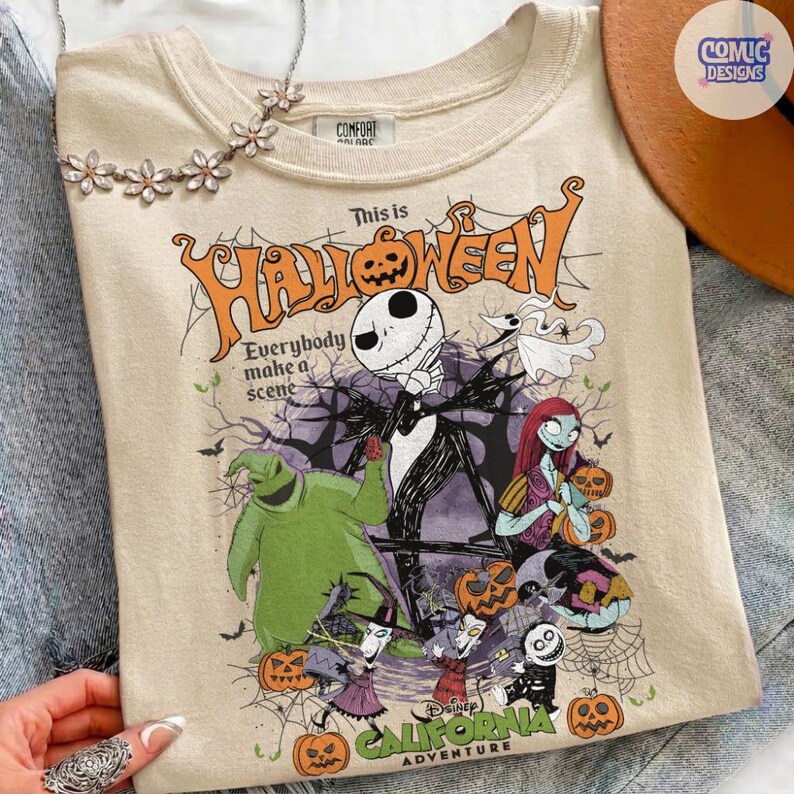 May include: Beige t-shirt featuring a Halloween-themed graphic with characters from The Nightmare Before Christmas. The design includes Jack Skellington, Sally, and Oogie Boogie, with the text "This is Halloween" and "California Adventure".