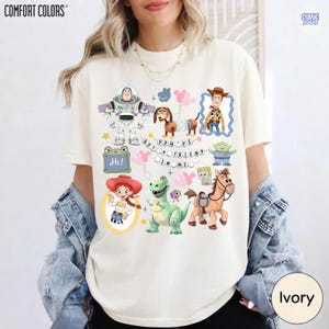 May include: An ivory t-shirt featuring a graphic print of various animated characters including a space ranger, a cowboy, a cowgirl, a dinosaur, a dog, and a horse. The text "YOU'VE GOT A FRIEND IN ME" is printed in the center with pink balloons.