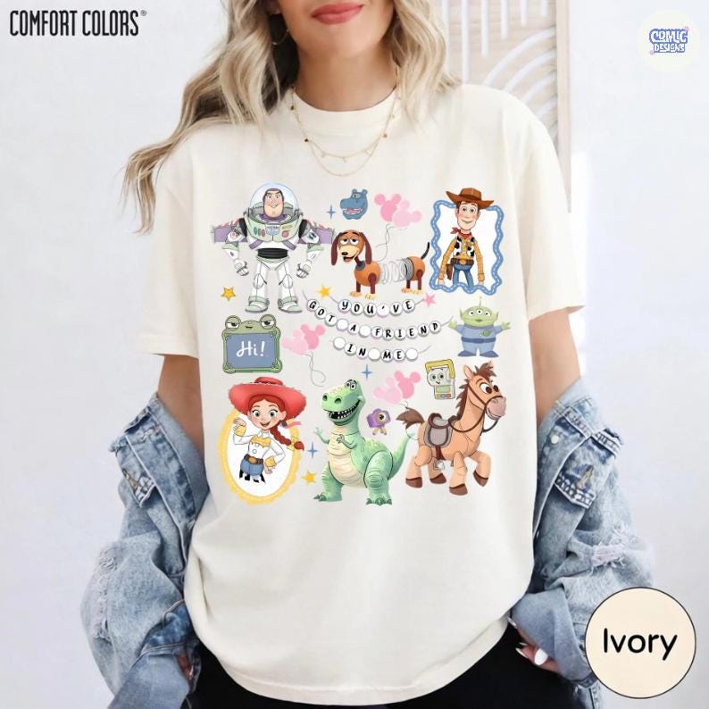 May include: An ivory t-shirt featuring a graphic print of various animated characters including a space ranger, a cowboy, a cowgirl, a dinosaur, a dog, and a horse. The text "YOU'VE GOT A FRIEND IN ME" is printed in the center with pink balloons.