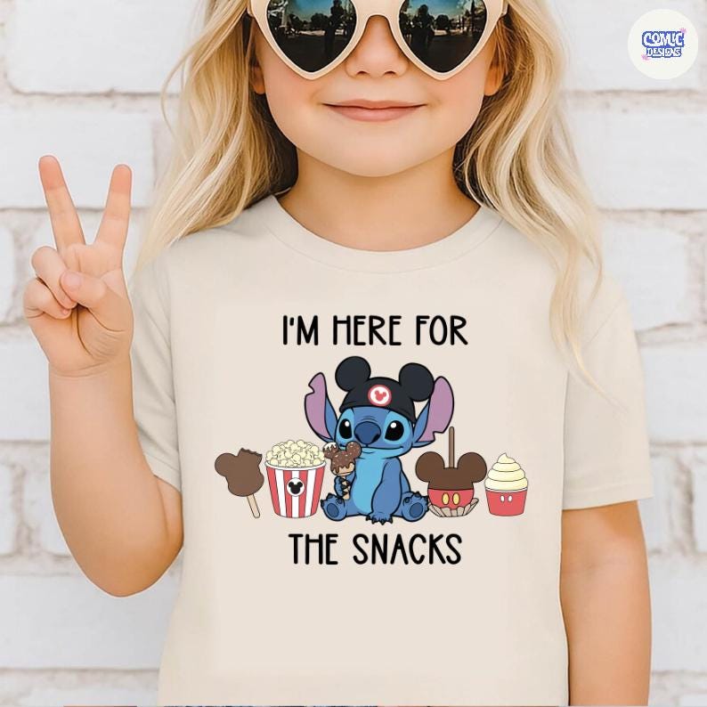May include: Beige t-shirt with the text "I'M HERE FOR THE SNACKS". The shirt features a blue cartoon character wearing a black hat, surrounded by popcorn, ice cream, and other treats. The child is wearing heart-shaped sunglasses.