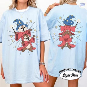 Puede incluir: Camiseta azul claro con una ilustraci&oacute;n de dibujos animados de dos ardillas listadas vestidas de magos, con sombreros azules y t&uacute;nicas rojas. La parte trasera de la camiseta muestra el mismo dise&ntilde;o. El texto "COMFORT COLORS Light Blue" est&aacute; impreso en la parte inferior.