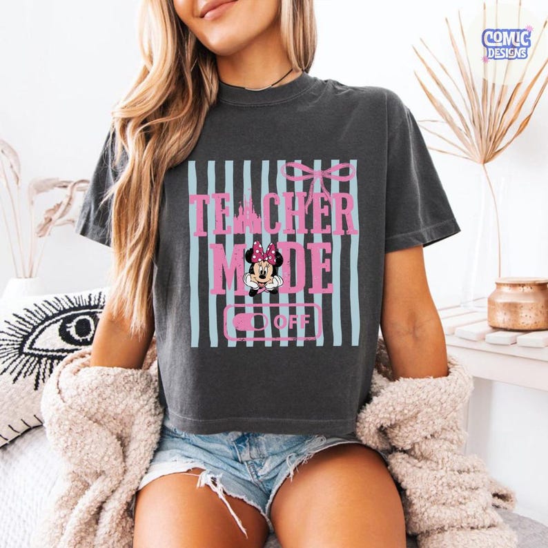 May include: Dark gray t-shirt with the text "TEACHER MODE OFF" in pink, with a Minnie Mouse graphic. The design includes light blue vertical stripes and a pink bow. The shirt is a casual, short-sleeved style.