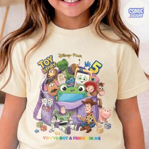 May include: A cream-coloured t-shirt featuring a colourful graphic of Toy Story characters including Woody, Buzz Lightyear, Rex, and Mr. Potato Head emerging from a purple backpack. The text reads Toy Story 5 and You've Got a Friend in Me.