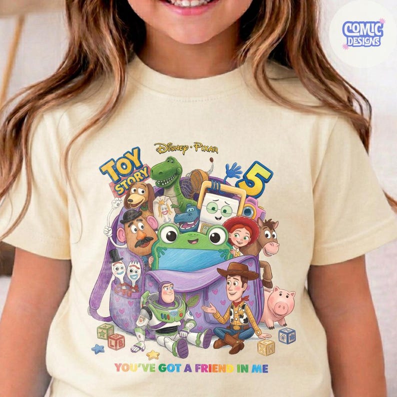 May include: A cream-coloured t-shirt featuring a colourful graphic of Toy Story characters including Woody, Buzz Lightyear, Rex, and Mr. Potato Head emerging from a purple backpack. The text reads Toy Story 5 and You've Got a Friend in Me.