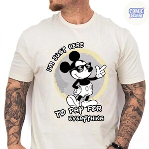May include: Cream-colored t-shirt featuring a cartoon mouse character wearing sunglasses with dollar signs. The text on the shirt reads "I'M JUST HERE TO PAY FOR EVERYTHING". The design is set against a yellow and gray circular background.