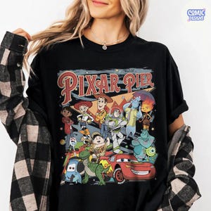Pu&ograve; includere: T-shirt nero con una grafica colorata di personaggi Pixar e la scritta "Pixar Pier". Il design include personaggi di vari film Pixar, come Woody, Buzz Lightyear e Saetta McQueen. La maglietta ha un classico girocollo.