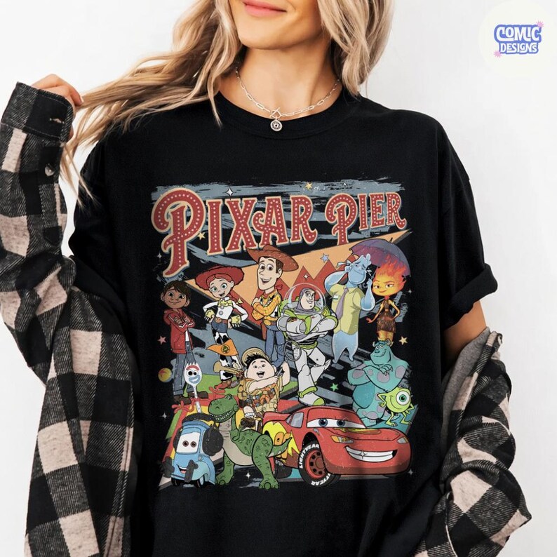 Pu&ograve; includere: T-shirt nero con una grafica colorata di personaggi Pixar e la scritta "Pixar Pier". Il design include personaggi di vari film Pixar, come Woody, Buzz Lightyear e Saetta McQueen. La maglietta ha un classico girocollo.