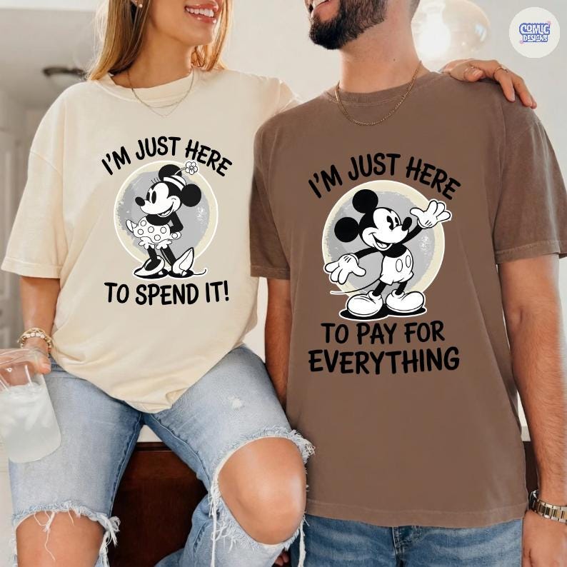 May include: Two t-shirts, one beige and one brown, featuring Mickey Mouse designs. The beige shirt has Minnie Mouse with the text "I'M JUST HERE TO SPEND IT!". The brown shirt has Mickey Mouse with the text "I'M JUST HERE TO PAY FOR EVERYTHING".