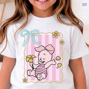 May include: White t-shirt featuring a cartoon illustration of Piglet from Winnie the Pooh. The design includes a pink and white striped background, a light blue bow, and yellow flowers. The shirt has a Comic Designs logo.