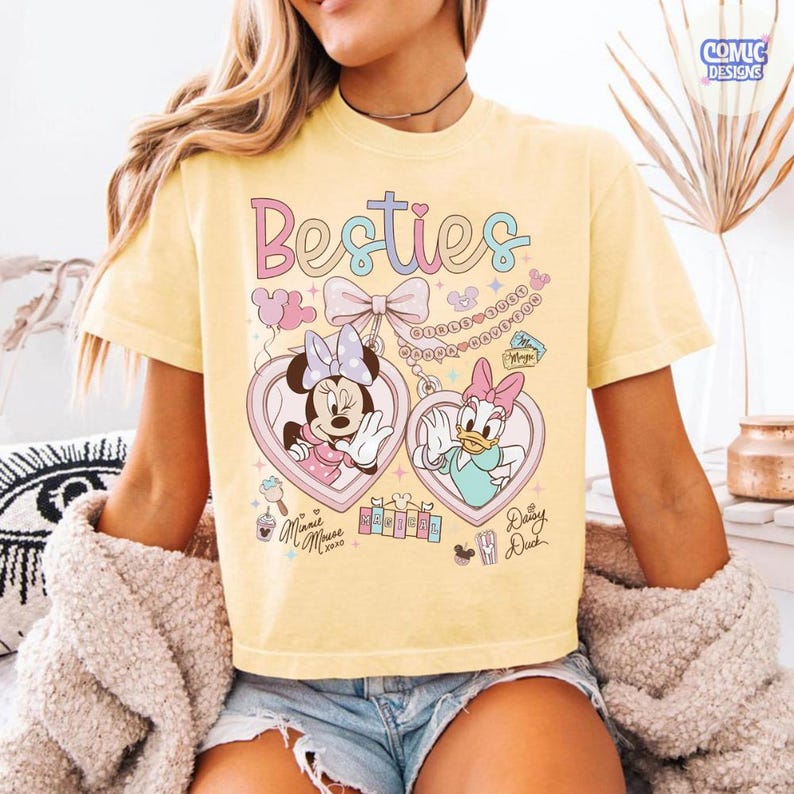 May include: Yellow t-shirt with the word "Besties" in pastel colors. The shirt features Minnie Mouse and Daisy Duck in heart-shaped lockets. The design includes bows, balloons, and the text "Minnie Mouse 2020" and "Daisy Duck".