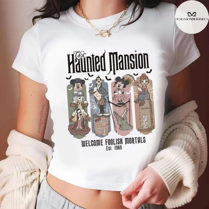 May include: White t-shirt featuring a graphic from The Haunted Mansion ride. The design includes the ride's name, cartoon characters, and the phrase "Welcome Foolish Mortals Est. 1969". The shirt has a crew neck and short sleeves.
