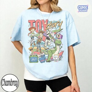 May include: A light blue t-shirt featuring a graphic print of Toy Story characters including Woody, Jessie, Buzz Lightyear, Rex, and Slinky Dog. The design includes the text Toy Story, Hi, and various colorful geometric shapes and star accents.