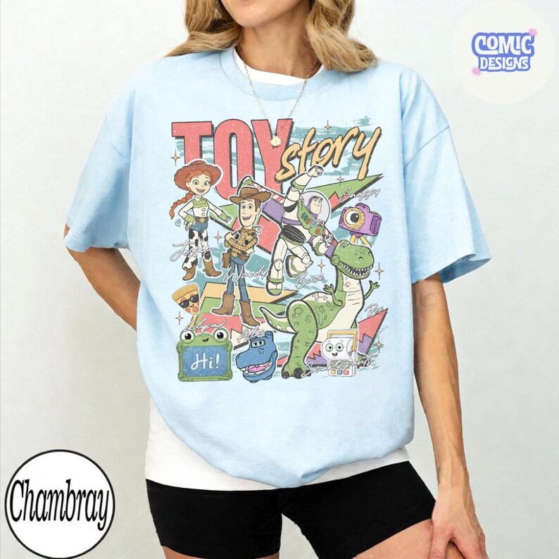 May include: A light blue t-shirt featuring a graphic print of Toy Story characters including Woody, Jessie, Buzz Lightyear, Rex, and Slinky Dog. The design includes the text Toy Story, Hi, and various colorful geometric shapes and star accents.