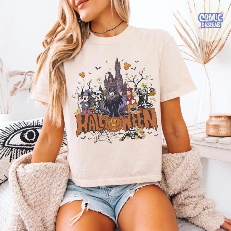 Puede incluir: Una camiseta de color crema con un estampado gr&aacute;fico de personajes de dibujos animados con disfraces de Halloween frente a un castillo. El dise&ntilde;o incluye el texto "HALLOWEEN" en letras grandes naranjas, murci&eacute;lagos, una telara&ntilde;a y una calabaza.