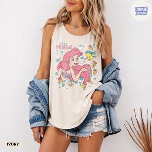 May include: An ivory tank top featuring a graphic print of Ariel from The Little Mermaid with pink hair and Flounder the fish, surrounded by blue bubbles. The text reads Disney The Little Mermaid. The tank top is worn with a blue denim jacket.