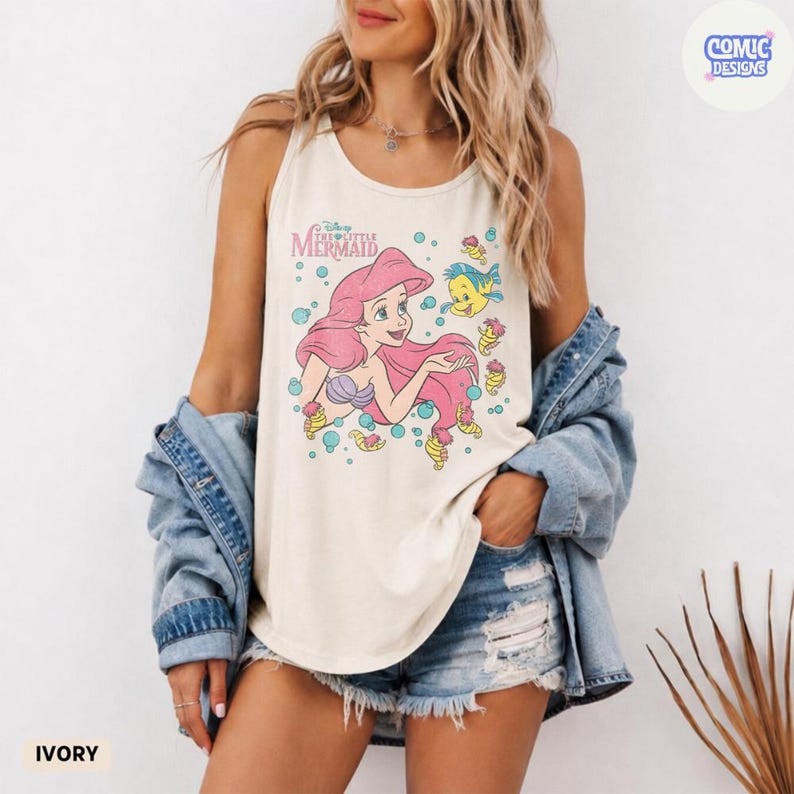 May include: An ivory tank top featuring a graphic print of Ariel from The Little Mermaid with pink hair and Flounder the fish, surrounded by blue bubbles. The text reads Disney The Little Mermaid. The tank top is worn with a blue denim jacket.