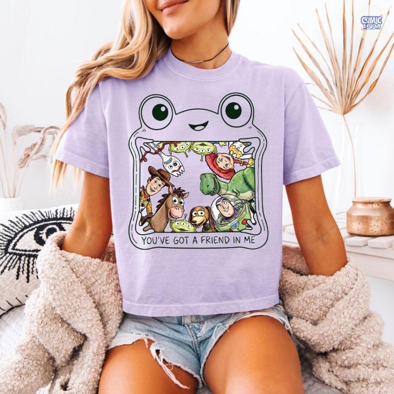 May include: A light purple t-shirt featuring a graphic of a frog outline containing various animated characters from the Toy Story franchise. The text "YOU'VE GOT A FRIEND IN ME" is printed in black capital letters below the character illustration.