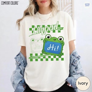 May include: An ivory t-shirt featuring a green checkered pattern and the word Lilypad in green script. A green frog character holds a blue sign with the text Hi! in white script, accompanied by a faint outline of a second frog character.