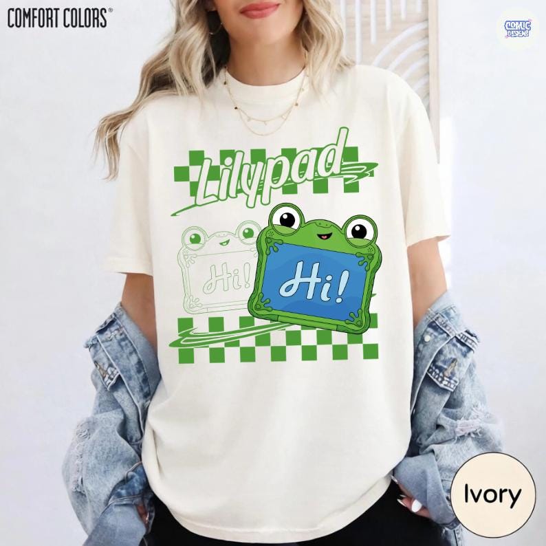 May include: An ivory t-shirt featuring a green checkered pattern and the word Lilypad in green script. A green frog character holds a blue sign with the text Hi! in white script, accompanied by a faint outline of a second frog character.