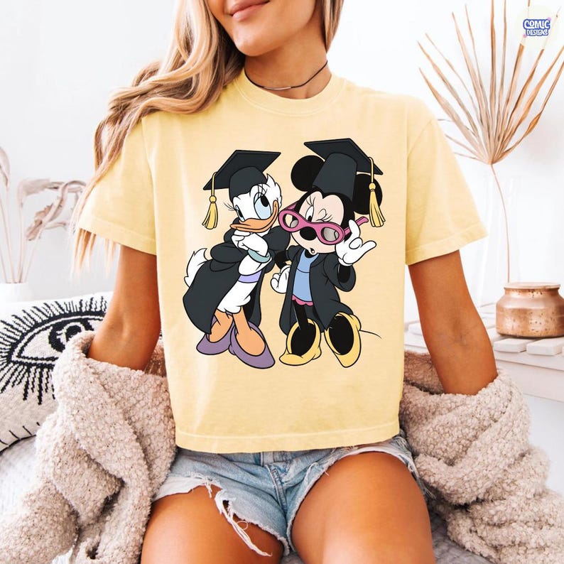 Puede incluir: Camiseta amarilla con un gr&aacute;fico de Daisy y Minnie Mouse con atuendo de graduaci&oacute;n. Los personajes de dibujos animados llevan vestidos y gorras de graduaci&oacute;n negros con borlas doradas. Minnie Mouse lleva gafas rosas.