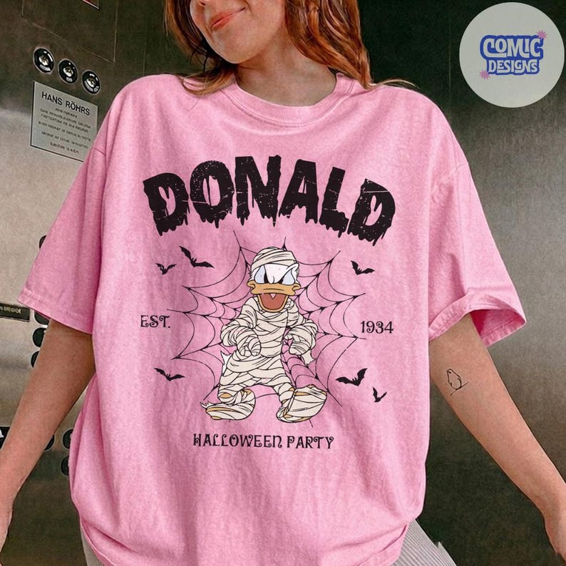 May include: A light pink t-shirt featuring a graphic of a cartoon duck wrapped in mummy bandages standing in front of a spider web. The text reads DONALD, EST. 1934, and HALLOWEEN PARTY, accompanied by small black bat silhouettes.