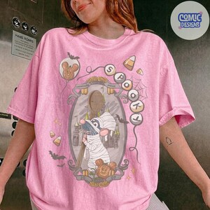 Puede incluir: Una camiseta rosa claro con un gr&aacute;fico de un rat&oacute;n vestido de momia sosteniendo una cuchara de madera. El dise&ntilde;o incluye un castillo, murci&eacute;lagos, dulces, telara&ntilde;as y el texto "SPOOKY" y "2007" dentro de un marco ovalado y globos.