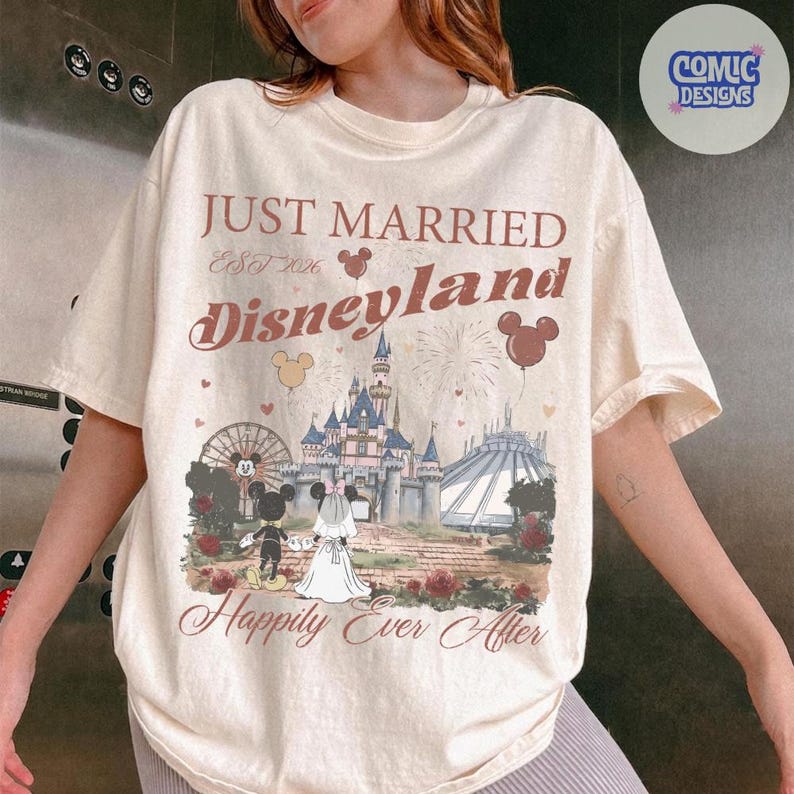 May include: Cream-colored t-shirt with a Disneyland-themed graphic. The design includes the text "Just Married Disneyland Est. 2026" and "Happily Ever After." The graphic features Mickey and Minnie Mouse, a castle, fireworks, and a Ferris wheel.