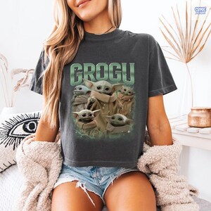 May include: Dark gray t-shirt featuring the word "GROGU" in green block letters above a graphic of Grogu from The Mandalorian. The graphic shows Grogu in various poses. The shirt is made of a soft material.