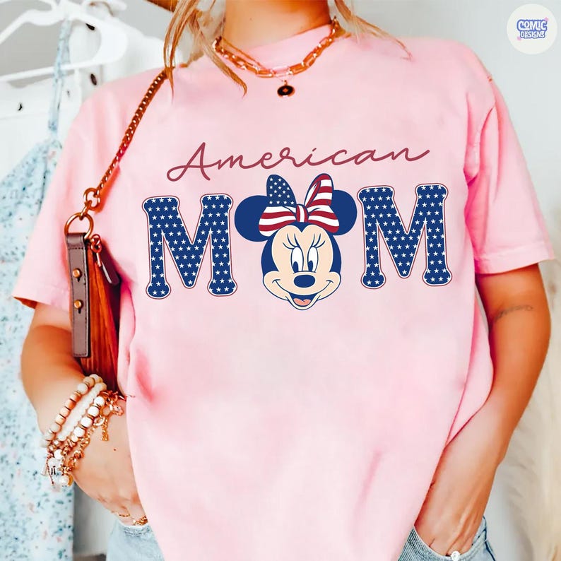 May include: Pink t-shirt with the words "American MOM" in a script font and block letters. The letter "M" features a Minnie Mouse graphic with a red, white, and blue bow. The shirt is a casual style.