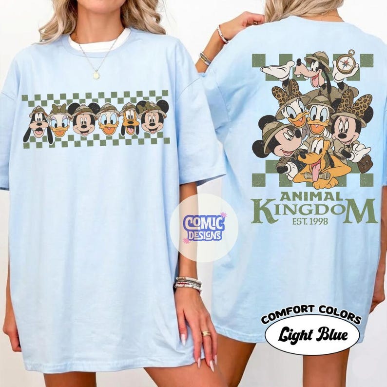 Pode incluir: Camiseta azul claro com um design de desenho animado de personagens da Disney com chap&eacute;us de safari. A frente da camisa mostra uma fileira de personagens, enquanto a parte de tr&aacute;s mostra um grupo maior com o texto "ANIMAL KINGDOM EST. 1998".