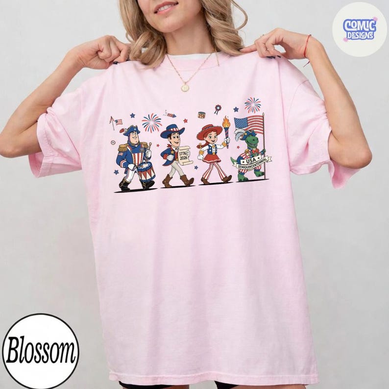 May include: A light pink t-shirt featuring a patriotic design with cartoon characters marching. The design includes a drummer, a cowboy, a cowgirl, and a dinosaur holding a flag and torch. Fireworks and flags are also included.