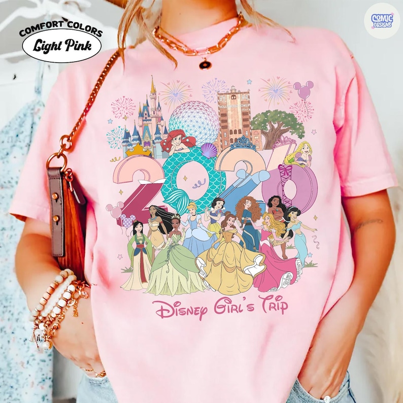 May include: A light pink t-shirt featuring a graphic print of the year 2024 with Disney characters, castle, and park icons. The text 'Disney Girl's Trip' is printed at the bottom. The shirt has a crew neck and short sleeves, with a Comfort Colors label.
