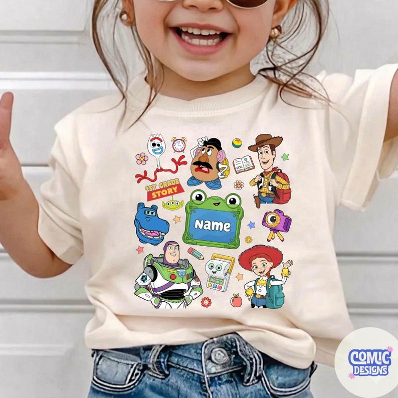 May include: A cream-colored short-sleeve t-shirt featuring a colorful graphic print of various cartoon characters including a cowboy, a space ranger, a cowgirl, a potato figure, and a fork character, with a green frame containing the text Name.