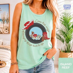May include: A light mint green sleeveless tank top featuring a graphic print of a tire frame containing cartoon characters, a red cowboy hat, and a red cowboy boot. The text reads, Love has ways of bringing things back to life.