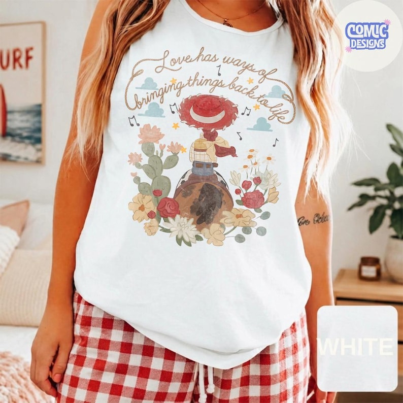 Op de afbeelding: Een wit mouwloos hemd met een grafische print van een cowgirl-figuur met een rode hoed, zittend op een bruin paard. Het ontwerp bevat kleurrijke bloemen, cactussen en de tekst "Love has ways of bringing things back to life."