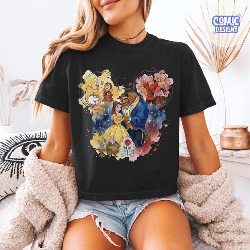 May include: A black short-sleeve t-shirt featuring a colourful graphic print of characters from Beauty and the Beast arranged in a Mickey Mouse head silhouette. The design includes Belle, the Beast, Gaston, and other characters with a rose.