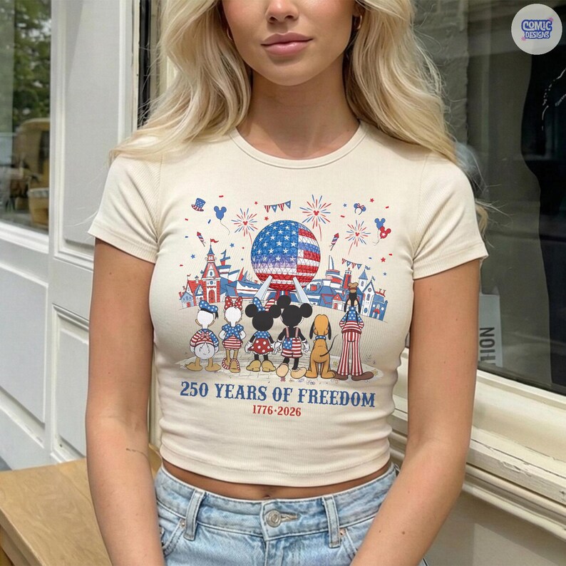 May include: A cream-colored short-sleeve t-shirt featuring a graphic of cartoon characters standing before a large sphere and fireworks. The text reads 250 YEARS OF FREEDOM 1776-2026. The design includes red, white, and blue patriotic elements.