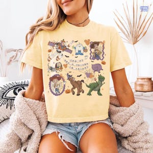 May include: A pale yellow short-sleeve t-shirt featuring a graphic print of various cartoon characters in Halloween costumes. The text "YOU'VE GOT A FRIEND IN FRIGHT" is centered among illustrations of a space ranger, a cowboy, a cowgirl, and a dinosaur.