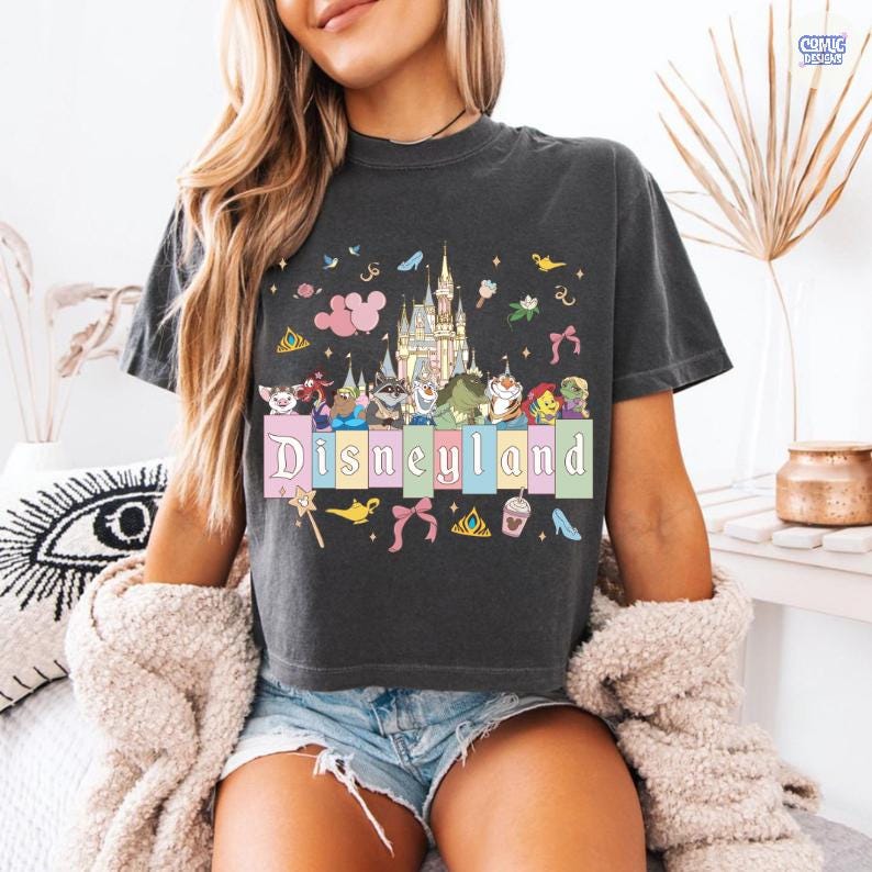 Pu&ograve; includere: T-shirt grigio scuro con grafica colorata di Disneyland. Il design include un castello, vari personaggi dei cartoni animati e la scritta "Disneyland" in colori pastello. Sono presenti anche elementi decorativi aggiuntivi come fiocchi, scarpe e corone.