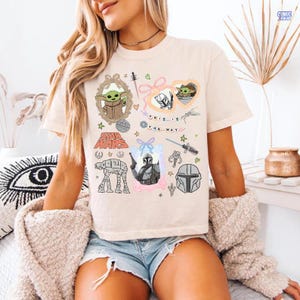 Puede incluir: Camiseta beige con gr&aacute;ficos de Star Wars, incluyendo el texto "THIS IS THE WAY" e im&aacute;genes de personajes como Grogu y el Mandaloriano. El dise&ntilde;o incorpora varios elementos del universo Star Wars.