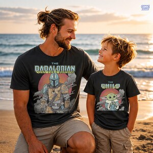 Puede incluir: Dos camisetas negras con gr&aacute;ficos a juego. Una camiseta dice "The Dadalorian" con un gr&aacute;fico de Mandaloriano. La otra dice "The Child" con un gr&aacute;fico de Grogu. Ambas camisetas tienen un dise&ntilde;o retro con un fondo de puesta de sol.
