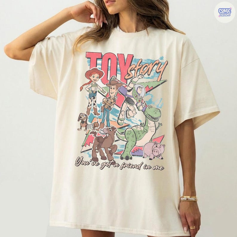 Może przedstawiać: Kremowy t-shirt z kolorową grafiką postaci z Toy Story. Projekt zawiera Woody'ego, Buzz'a Astrala, Jessie, Rexa i innych, ze słowami "Toy Story" i "You've got a friend in me".