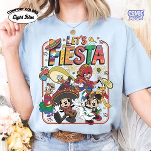 May include: Light blue t-shirt with a colorful graphic that says "Let's Fiesta". The graphic features cartoon characters in festive attire, including sombreros and musical instruments. The shirt has the words "Comfort Colors" and "Light Blue" printed on it.