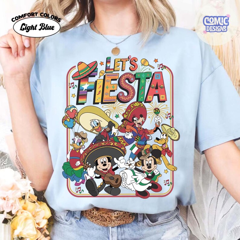 May include: Light blue t-shirt with a colorful graphic that says "Let's Fiesta". The graphic features cartoon characters in festive attire, including sombreros and musical instruments. The shirt has the words "Comfort Colors" and "Light Blue" printed on it.
