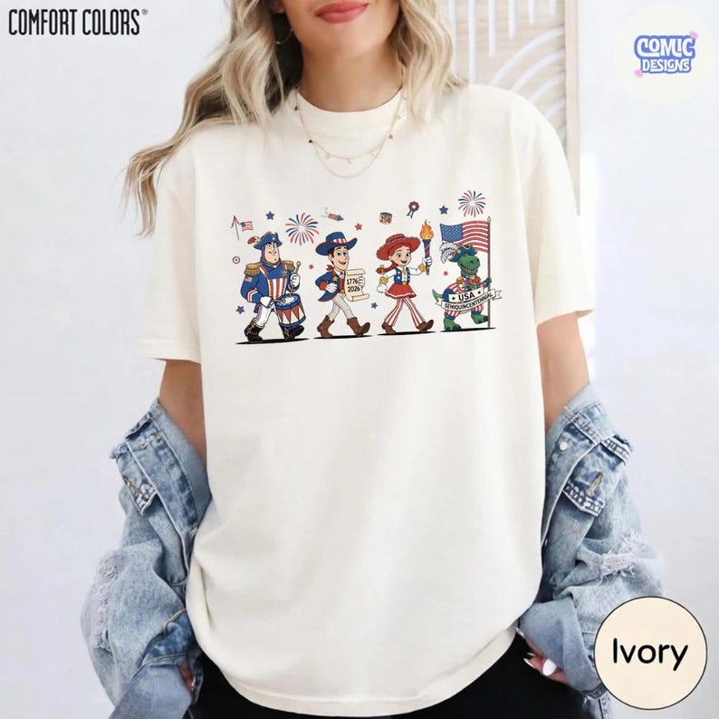 May include: Ivory-colored t-shirt featuring a patriotic cartoon parade with characters in red, white, and blue. The shirt has the words "COMFORT COLORS" and "COMIC DESIGNS" printed on it. The shirt is ivory in color.