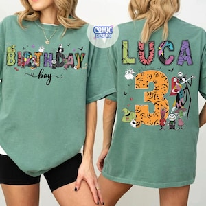 May include: A sage green t-shirt featuring a front graphic with the text Birthday boy and characters from The Nightmare Before Christmas. The back displays the name Luca, the number 3 in an orange pumpkin pattern, and various movie characters.